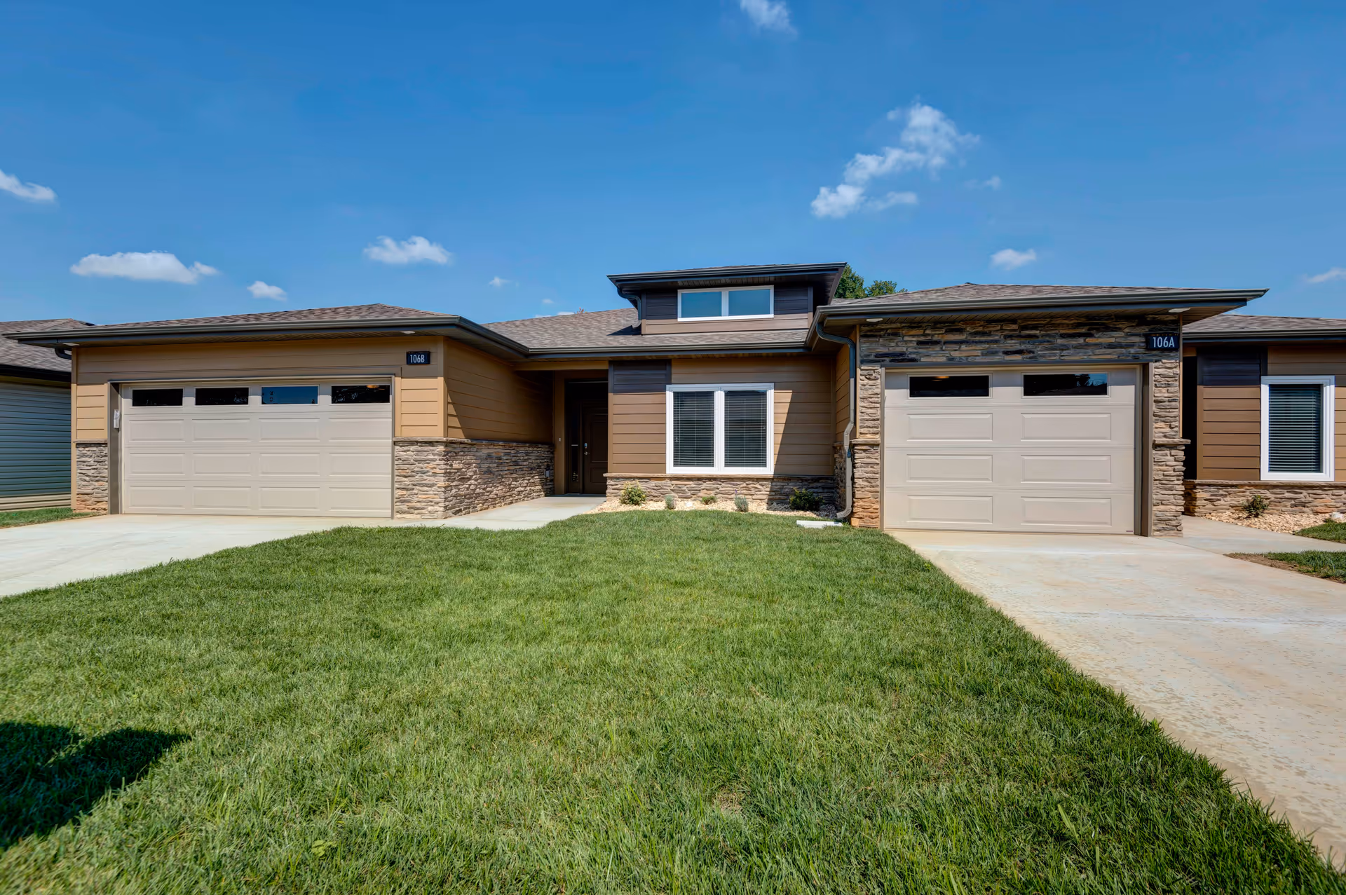Front exterior view of a single-story residential unit with two garages, a central entrance, stone and wood siding, and a well-maintained green lawn under a clear blue sky.