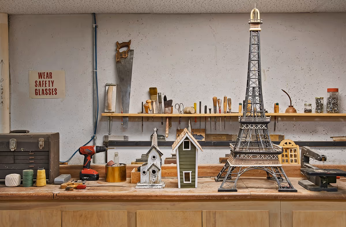 A workshop bench with various woodworking tools and materials including a saw, drill, paintbrushes, and jars of screws. On the bench are three wooden models: a small church, a house, and a large model of the Eiffel Tower. A sign on the wall reads 'WEAR SAFETY GLASSES'.