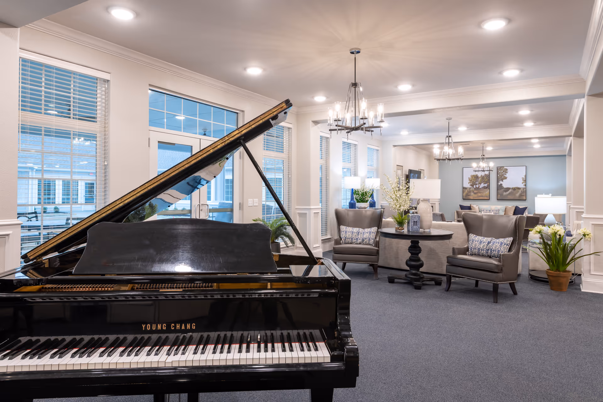 Common lounge featuring a black grand piano in the foreground and comfortable seating areas with armchairs, tables, and chandeliers.