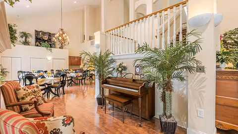 A bright and spacious interior common area featuring a wooden piano with a bench, two potted palm plants, and comfortable upholstered armchairs with floral patterns. In the background, there are multiple dining tables with black chairs and place settings, under a chandelier and high ceilings with white railings on the upper level.