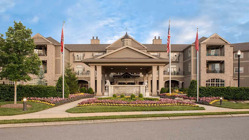 Front exterior view of Garden Plaza Of Greenbriar Cove, a large multi-story building with a covered entrance, surrounded by well-maintained landscaping including flower beds, shrubs, and trees. There are three flagpoles with flags in front of the entrance.