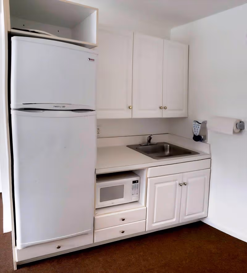 A small kitchen area with a white refrigerator, white cabinets, a microwave oven, a stainless steel sink, and a paper towel holder mounted on the wall.