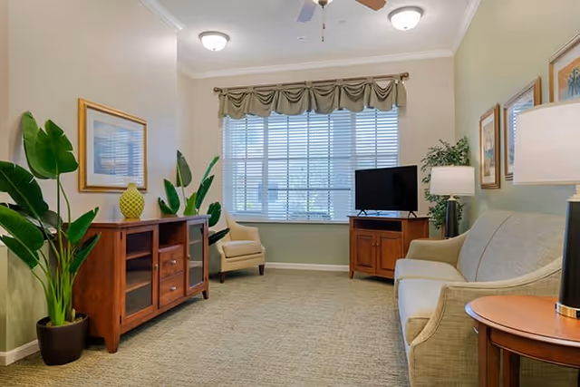 A cozy living room with a beige sofa, a wooden side table with a lamp, a wooden TV stand with a flat-screen TV, two beige armchairs, a wooden cabinet with glass doors, green potted plants, framed artwork on the walls, and a large window with blinds and a valance.