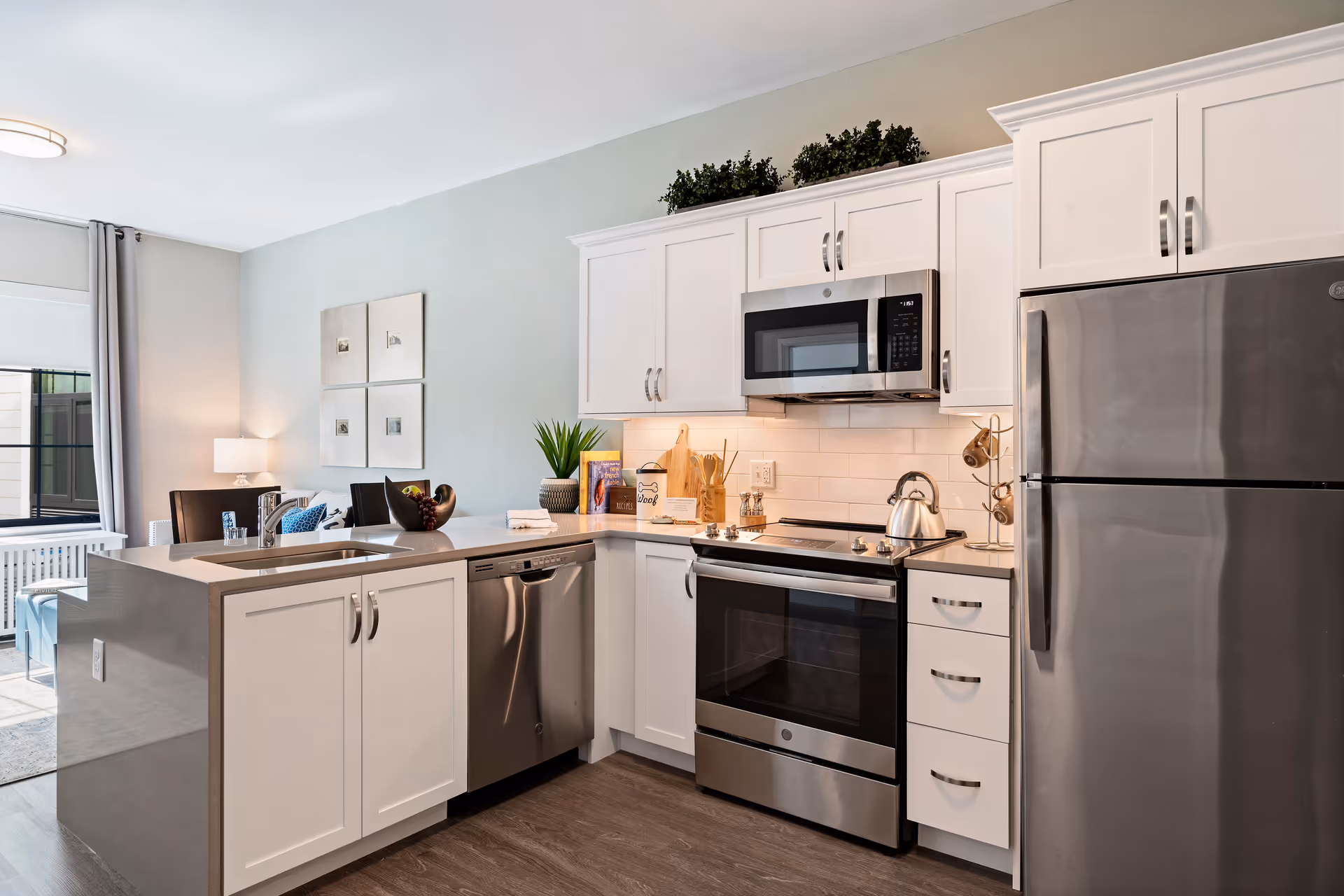 Modern kitchen with white cabinets, stainless steel appliances including a refrigerator, oven, microwave, and dishwasher. A kitchen island with a sink and faucet is in the foreground. The kitchen opens into a living area with a window, curtains, and a lamp.