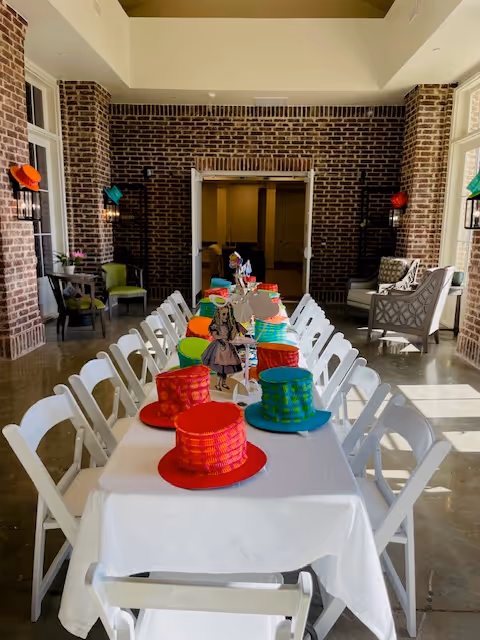 Long decorated dining table with white folding chairs and colorful hats in a brick-walled common room.