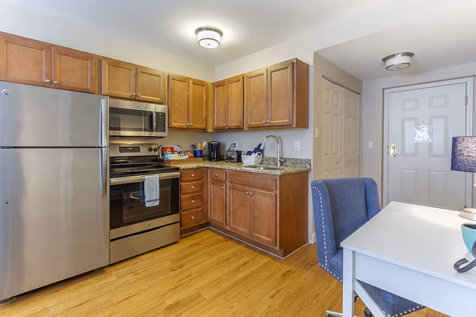 Compact kitchen with stainless steel refrigerator and oven, wooden cabinets, granite countertops, and a desk with a blue upholstered chair.