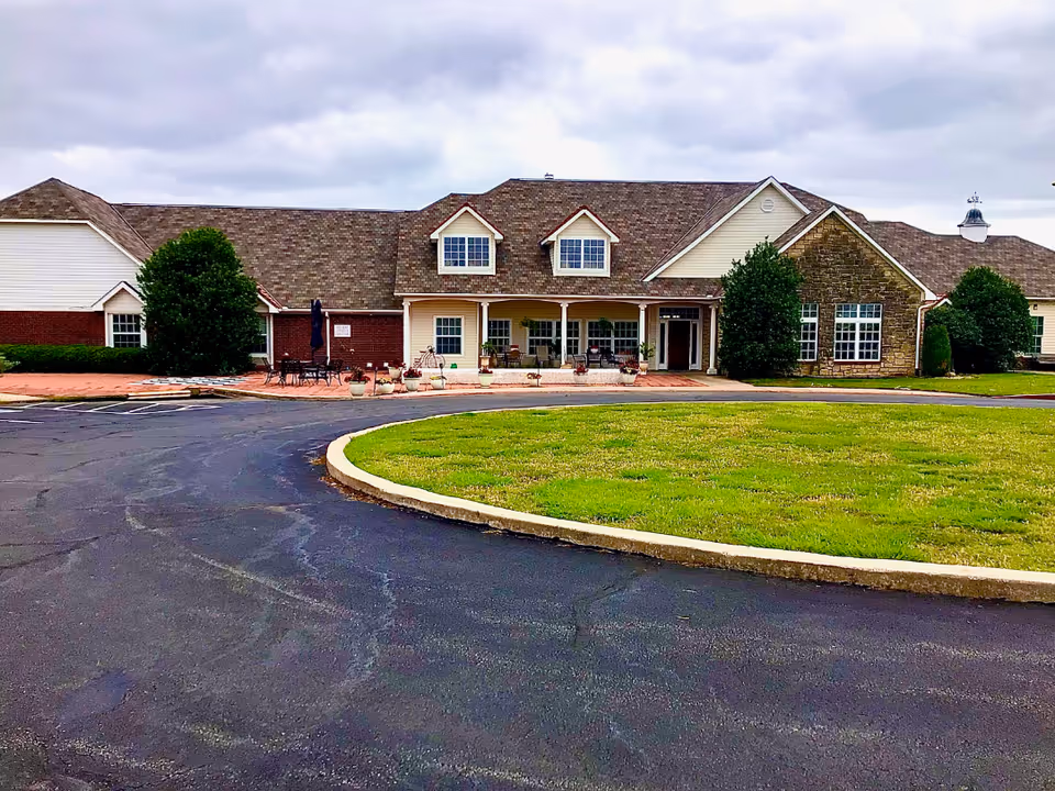 Front exterior of a single-story senior living building with a covered porch, patio seating, dormer windows, and a circular driveway with a grassy center.