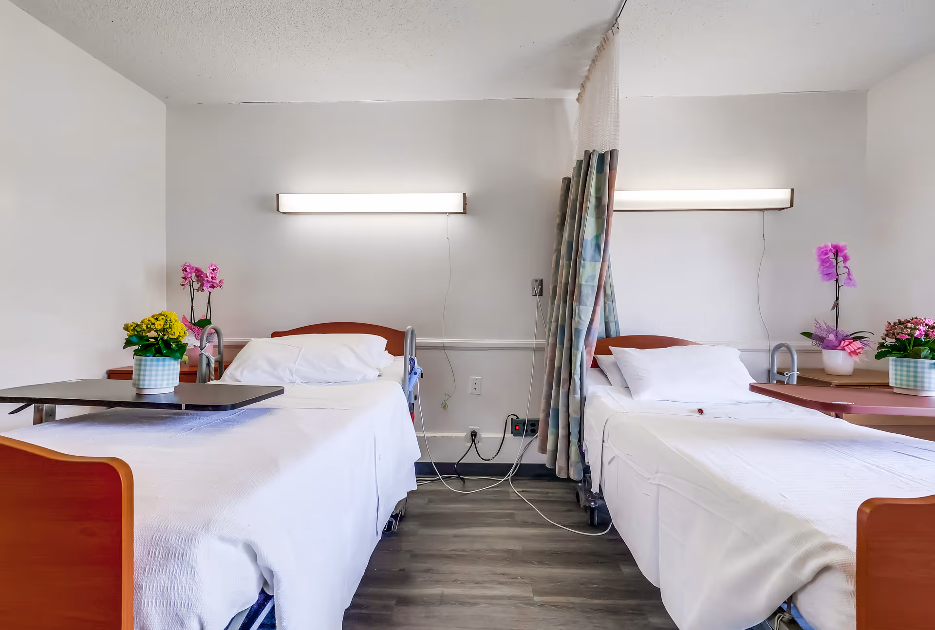 A hospital or post-acute care room with two single beds separated by a privacy curtain. Each bed has white bedding, a pillow, and a bedside table with colorful potted flowers. There are wall-mounted lights above each bed and medical equipment outlets on the wall behind the beds.