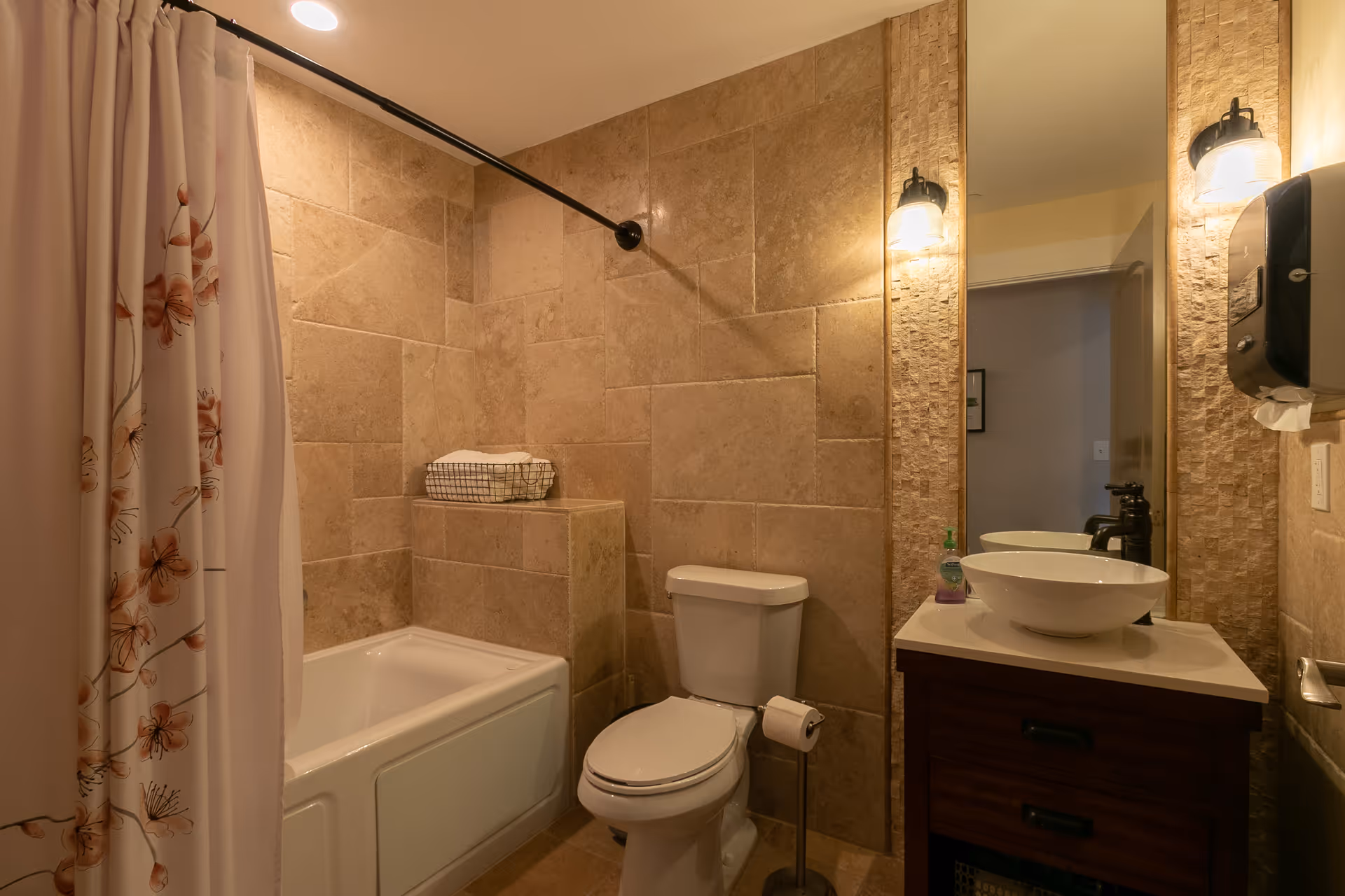 A bathroom with beige tiled walls and floor, featuring a white bathtub with a floral shower curtain, a white toilet with a toilet paper holder, and a dark wood vanity with a white vessel sink and a large mirror above it. Two wall-mounted lights flank the mirror, and a soap dispenser is on the sink countertop.