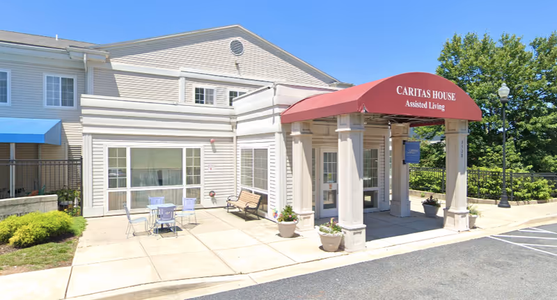 Exterior view of Caritas House Assisted Living facility showing the entrance with a red canopy displaying the facility name. There are potted plants near the entrance, a bench, and a small outdoor seating area with a table and chairs on the concrete patio. The building is light-colored with multiple windows and surrounded by greenery.