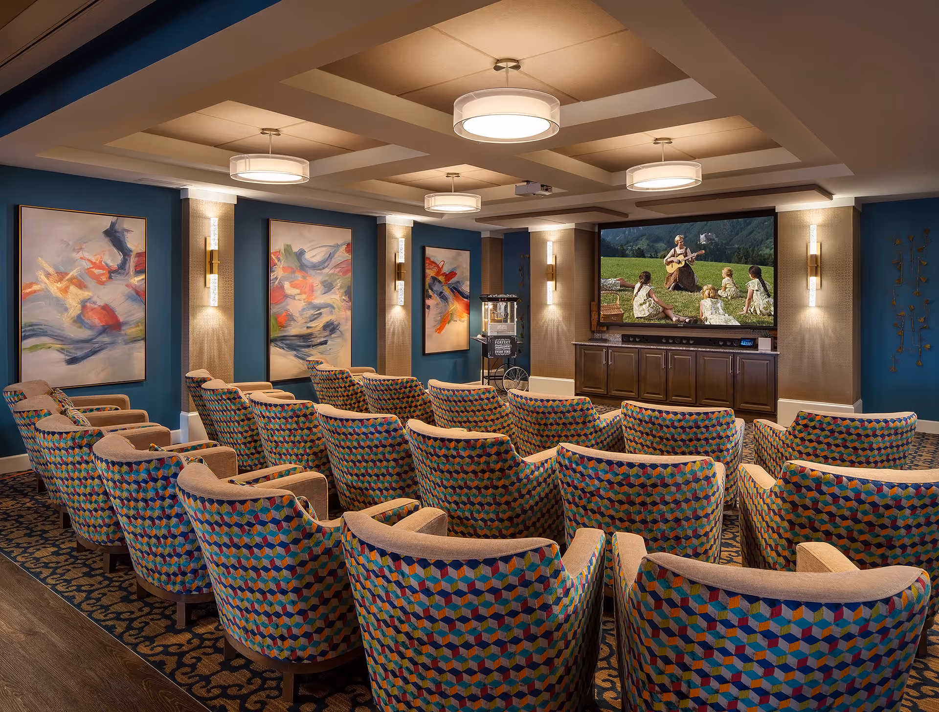 A cozy movie theater room with multiple rows of colorful patterned armchairs facing a large screen showing a woman playing guitar to children sitting on grass. The room has blue walls adorned with abstract paintings and modern wall sconces, with ceiling lights providing warm illumination.