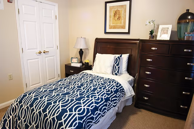 A neatly made single bed with a dark wooden headboard, covered with a blue and white patterned blanket and white pillows. Next to the bed is a dark wooden nightstand with a lamp, a small picture frame, and a flower vase. On the right side of the bed is a dark wooden dresser with multiple drawers, decorated with a framed photo, a small plant, and a large decorative vase. The walls are light-colored, and there is a framed artwork hanging above the bed. A closed white double-door closet is visible on the left side of the room.