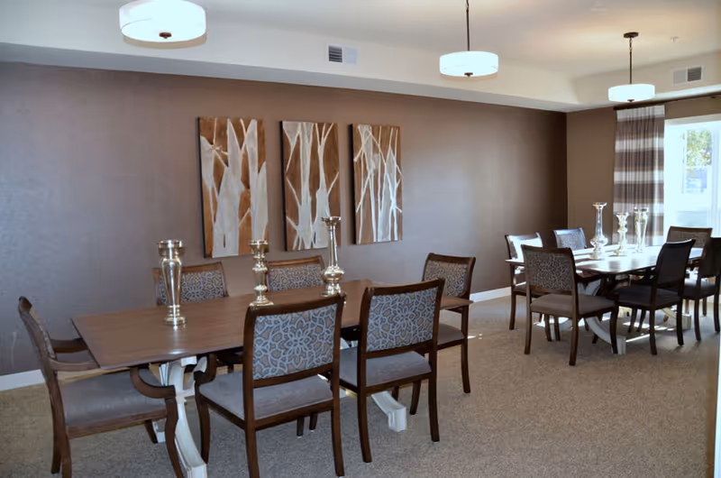 A dining room with two wooden tables, each surrounded by six chairs with patterned upholstery. The tables have decorative silver candle holders on them. The walls are painted brown and decorated with three vertical abstract paintings featuring tree designs. The room is well-lit with three ceiling lights and natural light coming through a window with striped curtains.