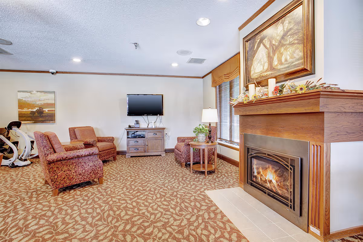 A cozy living room area with patterned carpet, three upholstered armchairs, a wooden side table with a lamp and plant, a flat-screen TV mounted on the wall above a wooden media cabinet, and a fireplace with a wooden mantle decorated with candles and flowers. There is a large framed painting above the fireplace and a window with blinds and a valance.