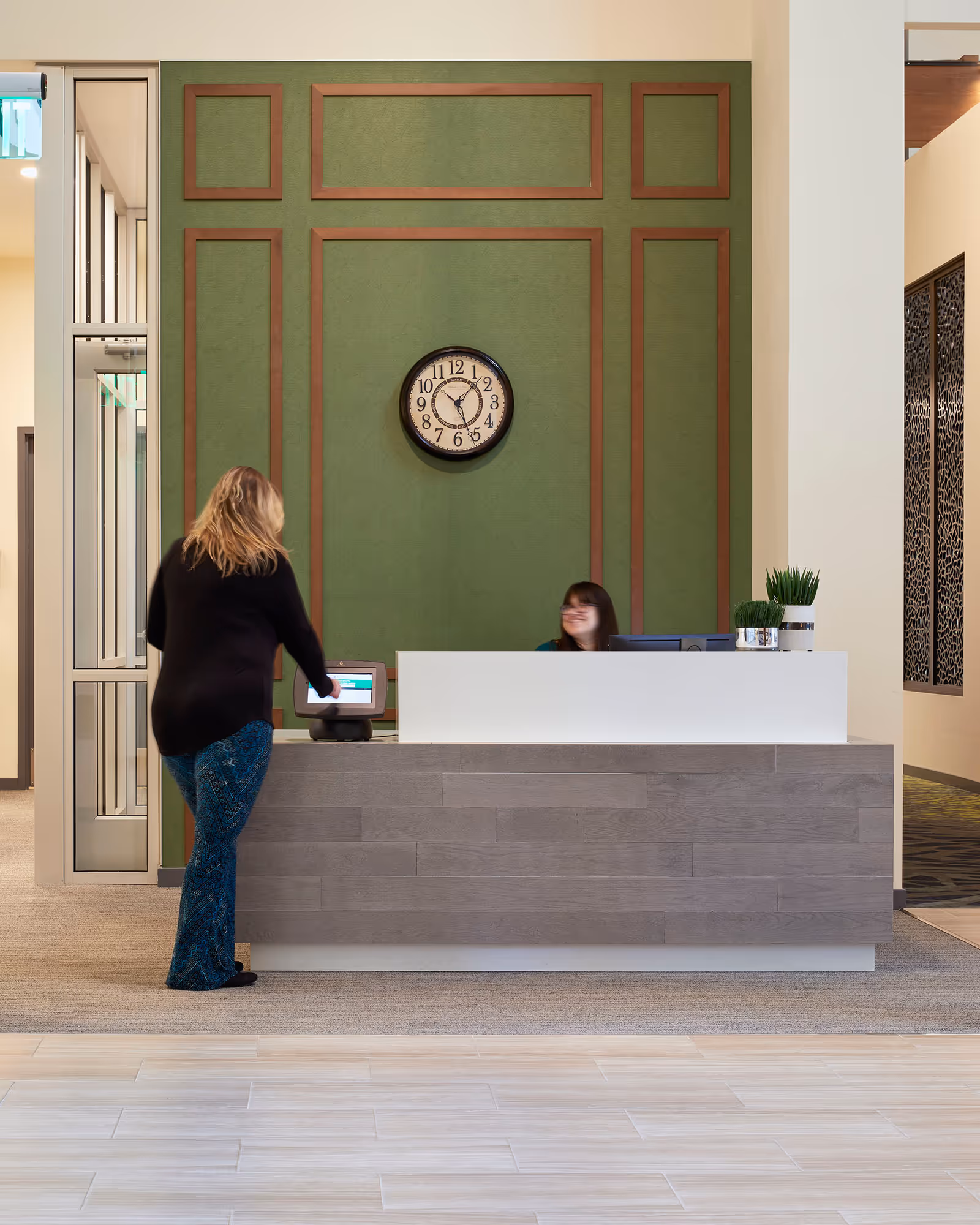 Reception area with a woman standing at the front desk interacting with a touchscreen device, and a receptionist smiling behind the desk. The wall behind the desk is green with wooden trim and a round clock showing the time as 5:25.