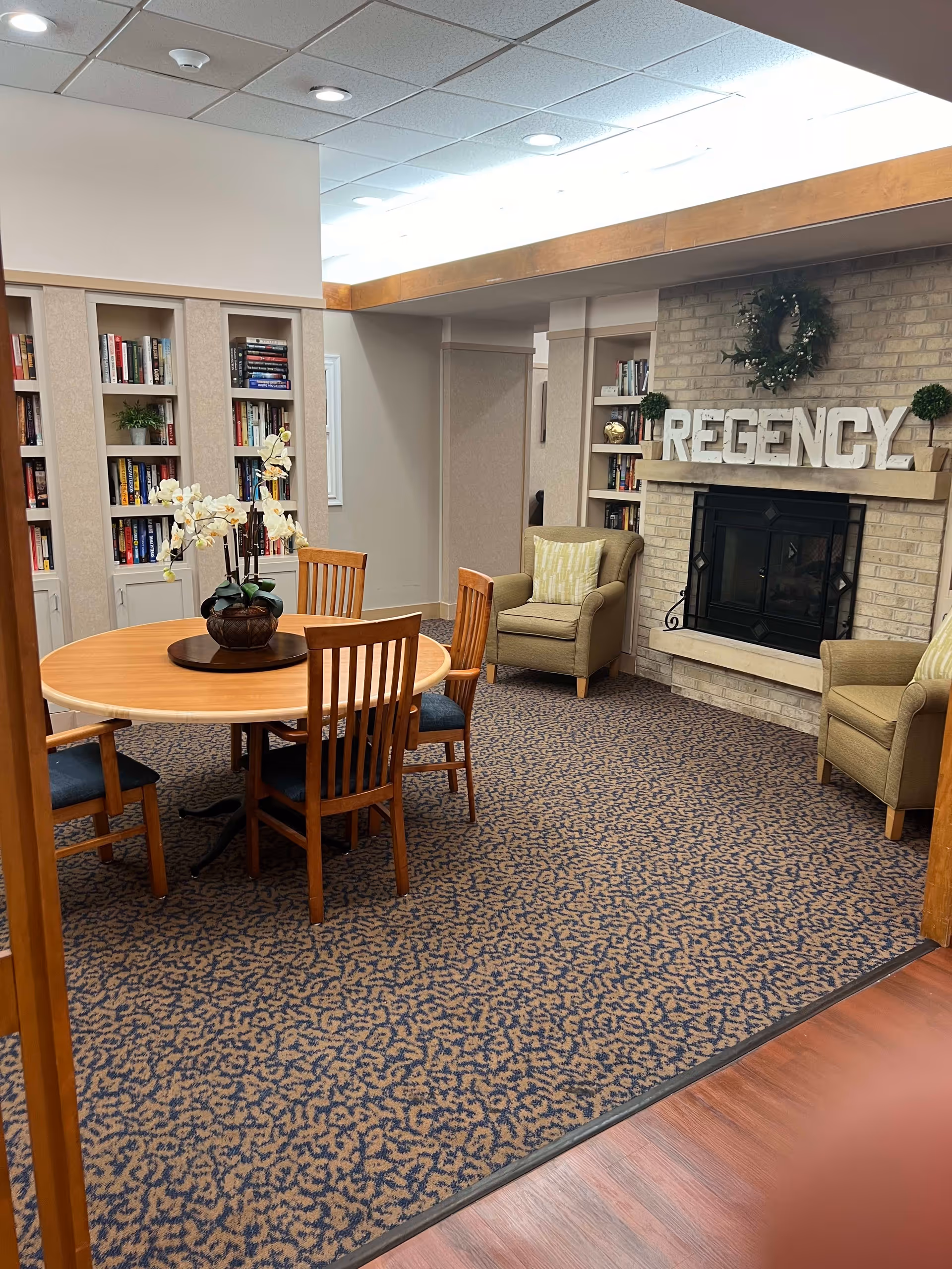 A cozy senior living community common area with a round wooden table surrounded by four wooden chairs with blue cushions. There are two beige armchairs with striped cushions near a brick fireplace. Above the fireplace, large white letters spell out 'REGENCY' and a green wreath is hung on the wall. Built-in bookshelves filled with books and decorative plants flank the fireplace. The room has patterned carpet and a ceiling with recessed lighting.