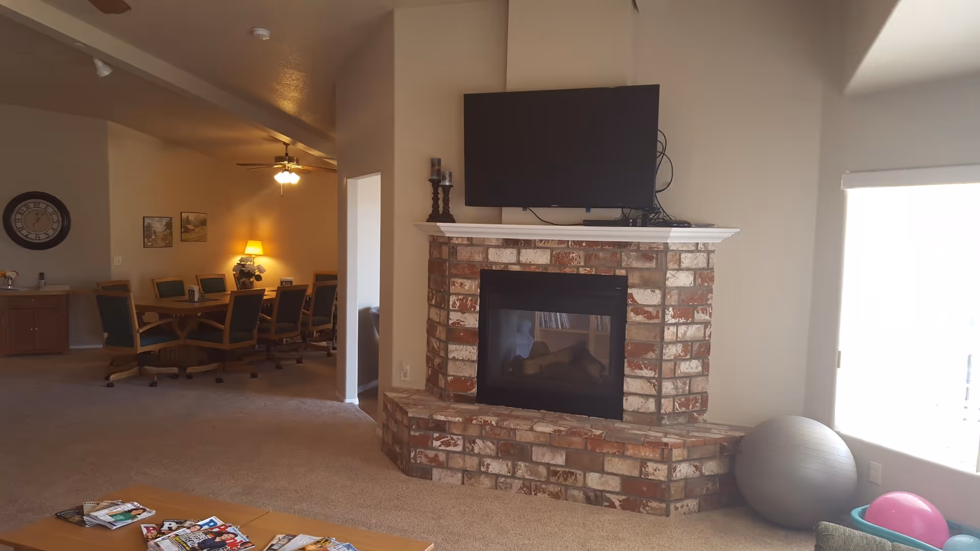 A cozy senior living common area featuring a brick fireplace with a flat-screen TV mounted above it. To the left, there is a dining table with several chairs, a ceiling fan with lights, a wall clock, and framed pictures on the wall. On the right side near a window, there is a large exercise ball and a pink ball in a basket. A coffee table with magazines is visible in the foreground.