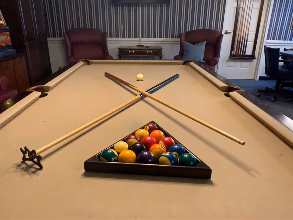 A billiards table with a beige felt surface, two crossed pool cues, a white cue ball, and a triangular rack filled with colorful billiard balls. In the background, there are two maroon armchairs, a small wooden table, and a wall with vertical striped wallpaper.