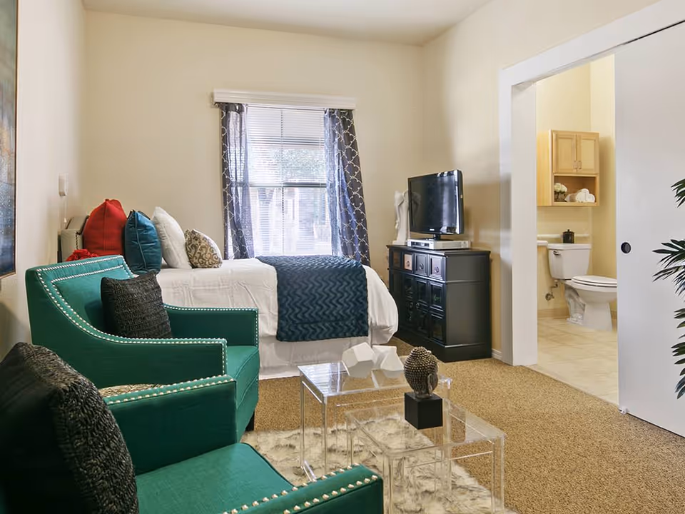 A cozy assisted living room with a bed adorned with multiple pillows and a blue throw blanket, two green armchairs with black cushions, a clear acrylic coffee table with decorative items, a window with patterned curtains, and a TV on a black cabinet. An open doorway reveals a bathroom with a toilet and wooden cabinet.