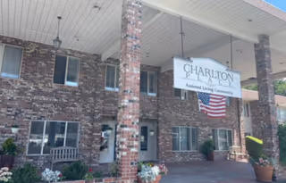 Entrance area of Charlton Place Assisted Living Community showing a brick building with a covered porch supported by brick columns. There is a hanging sign with the facility name and an American flag beneath it. Various potted plants and flowers are arranged near the entrance.