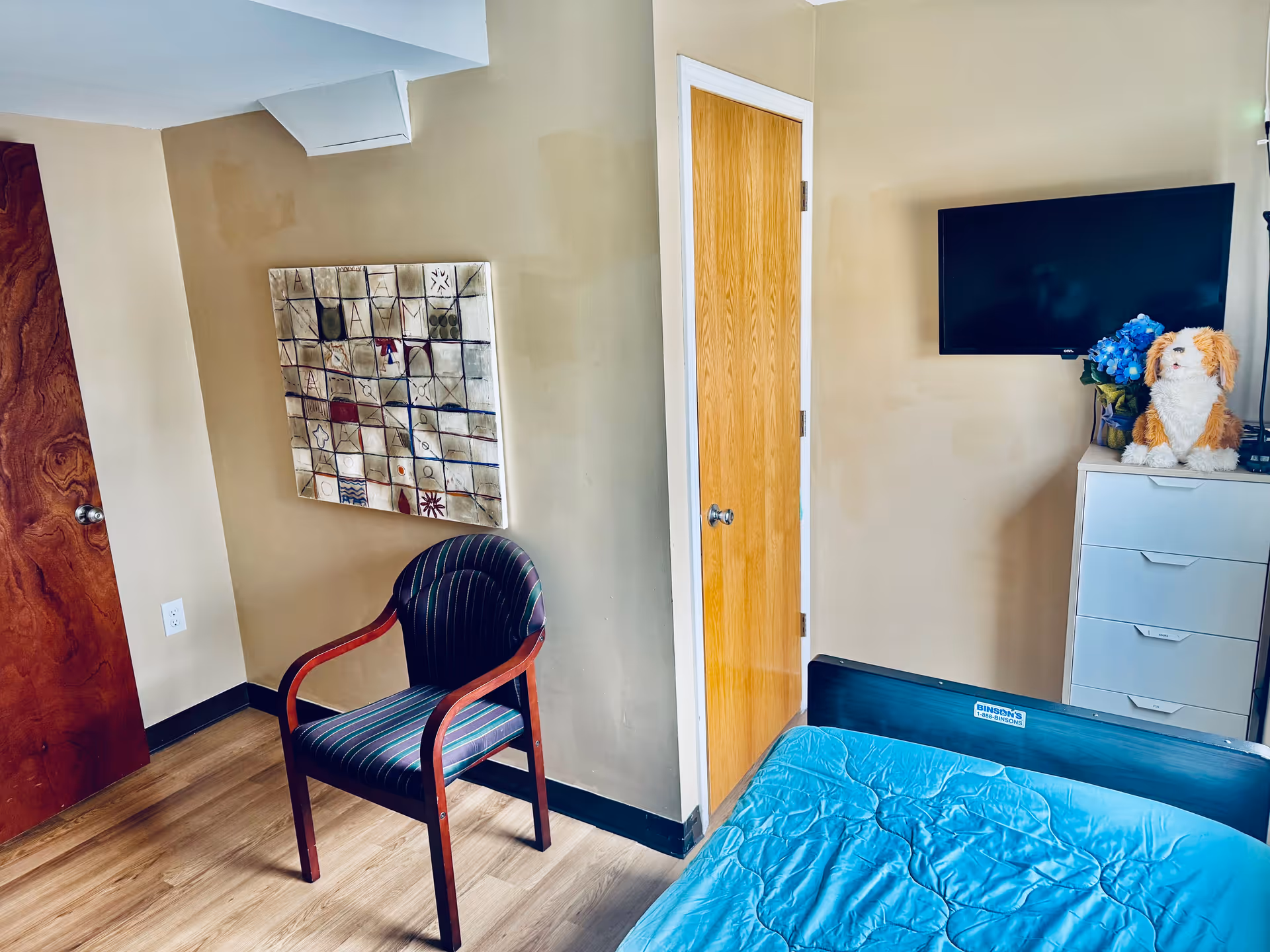 A small bedroom with a wooden door, a striped armchair, a bed with a blue quilt, a white dresser with a stuffed dog toy and a vase of blue flowers on top, and a wall-mounted flat screen TV.
