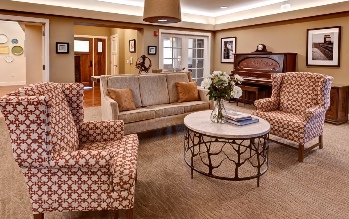 A cozy living room area with a beige sofa adorned with two brown pillows, two patterned armchairs in red and white, and a round coffee table with a decorative metal base and a vase of white flowers on top. In the background, there is a wooden piano with a clock on top, framed black and white photographs on the wall, and a set of French doors leading to another room. The room has warm beige walls and carpeted flooring.