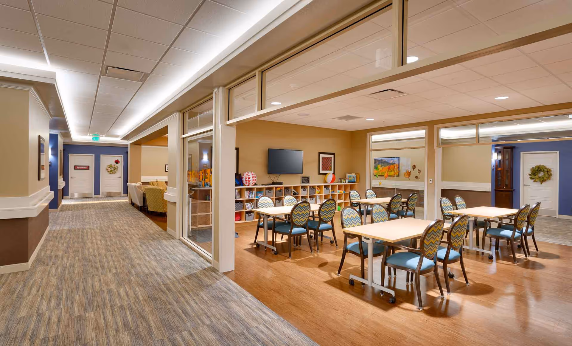 Well-lit communal dining and activity room with multiple tables and patterned chairs, shelving, a wall-mounted TV, and an adjacent hallway.