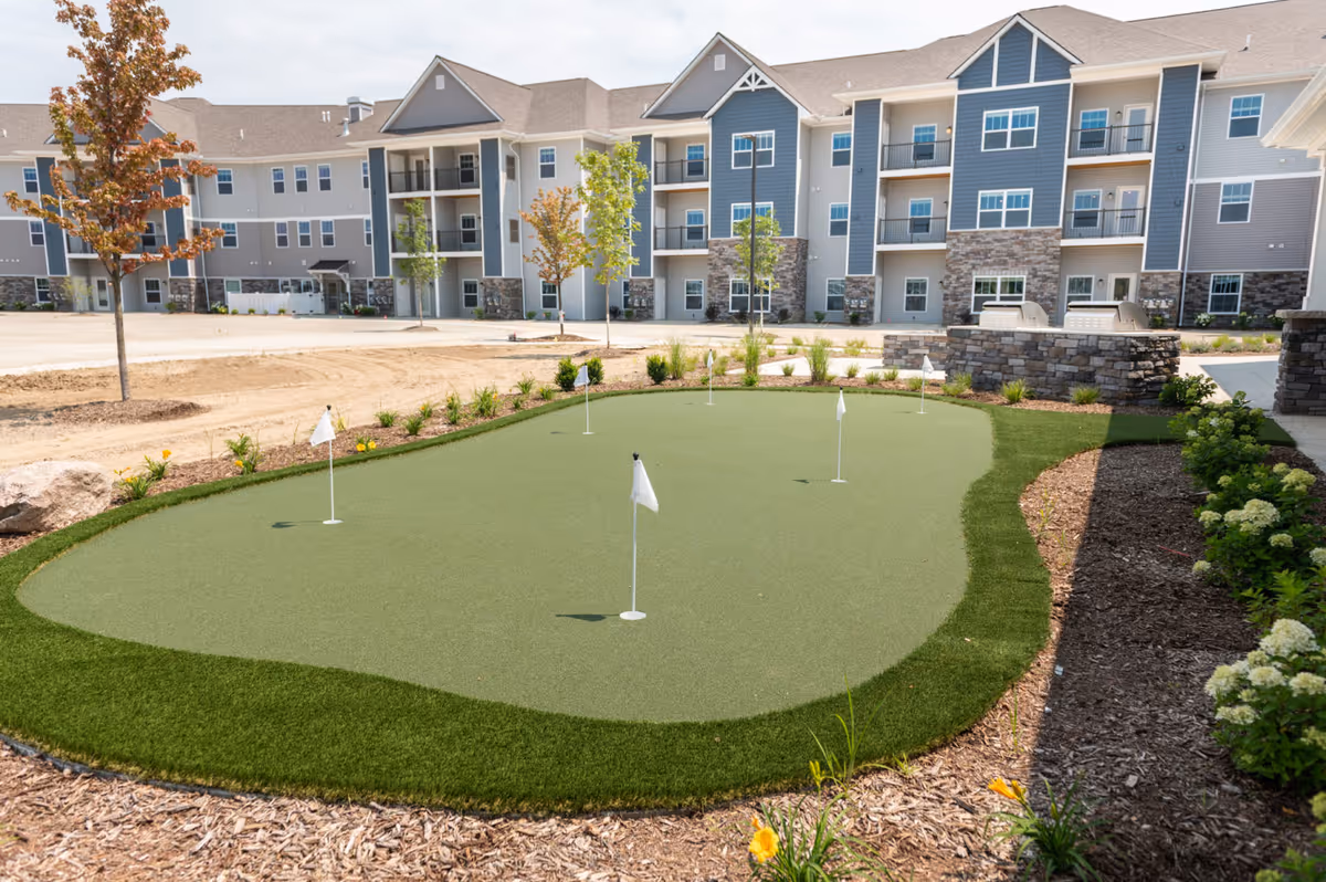 Outdoor putting green with five white flags surrounded by landscaping and mulch, with a multi-story residential building in the background under a partly cloudy sky.