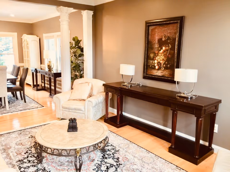 A well-appointed living room with an upholstered armchair, round marble coffee table, long dark console table with two lamps and framed art, and a dining area visible in the background.