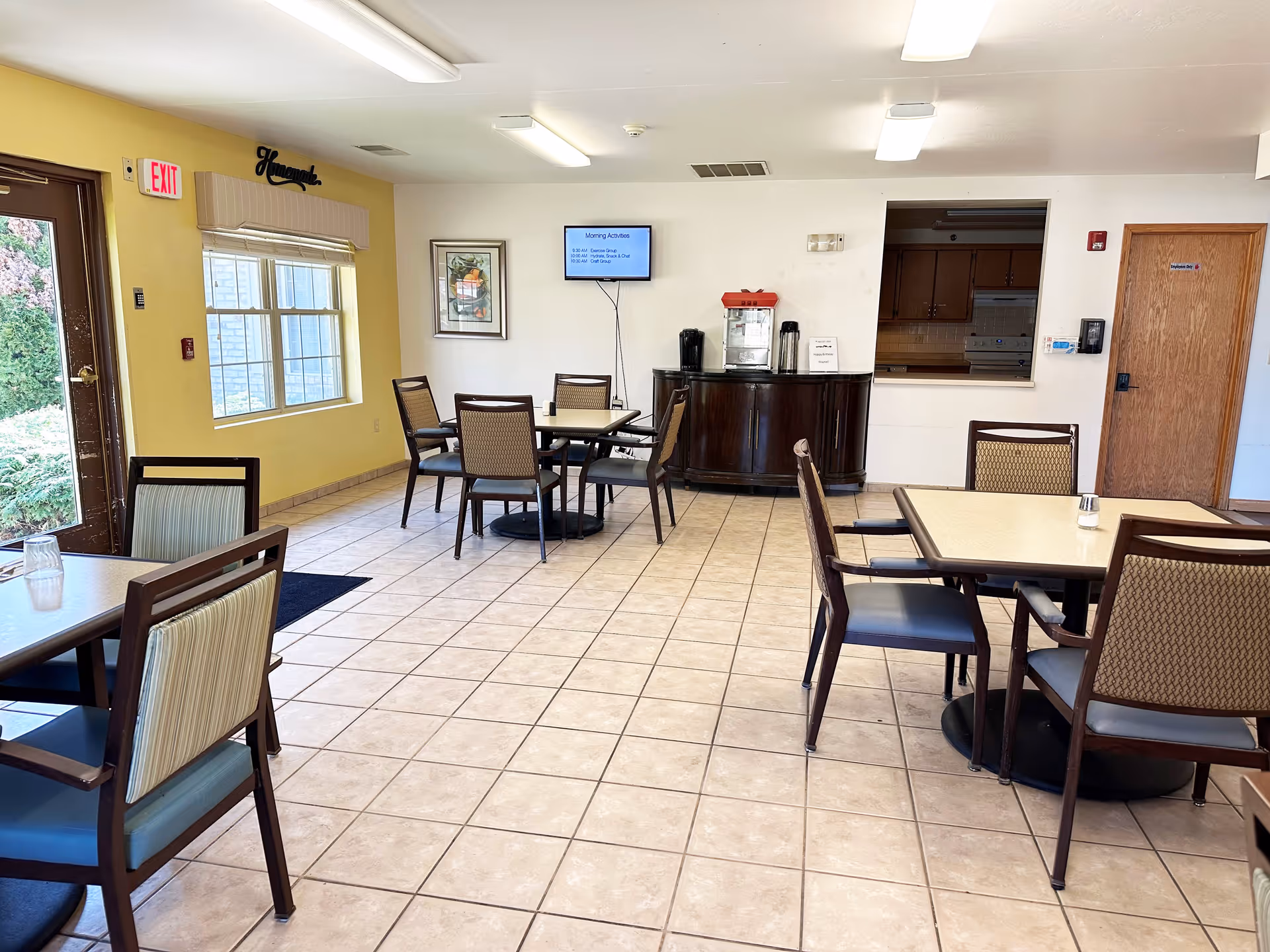 A dining area in a senior living facility with several tables and chairs arranged on a tiled floor. There is a window with blinds on the left wall, a door with an exit sign above it, and a small cabinet against the back wall holding a popcorn machine and coffee dispensers. A TV screen mounted on the back wall displays morning activities. The walls are painted yellow and white, and there is a pass-through window to a kitchen area on the right side.