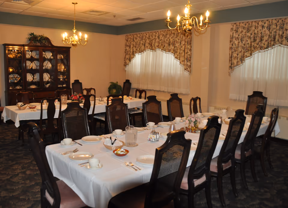 Formal dining room with long tables set for a meal, chandeliers overhead, wooden chairs, and a china cabinet.
