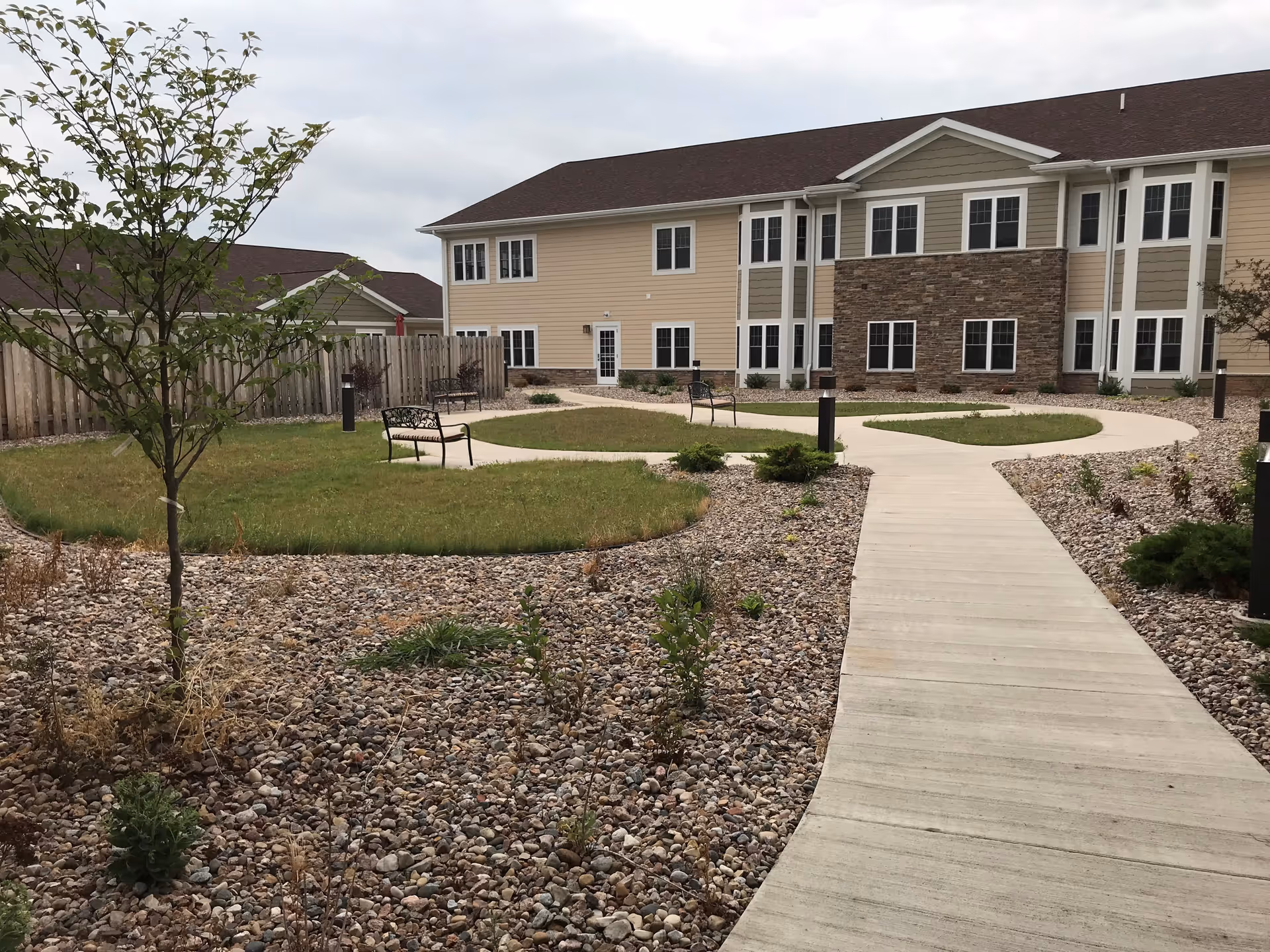 Outdoor courtyard area at Oak Park Place Green Bay featuring a concrete walkway, small grassy patches, benches, young trees, and a two-story building with beige and stone exterior in the background under a cloudy sky.