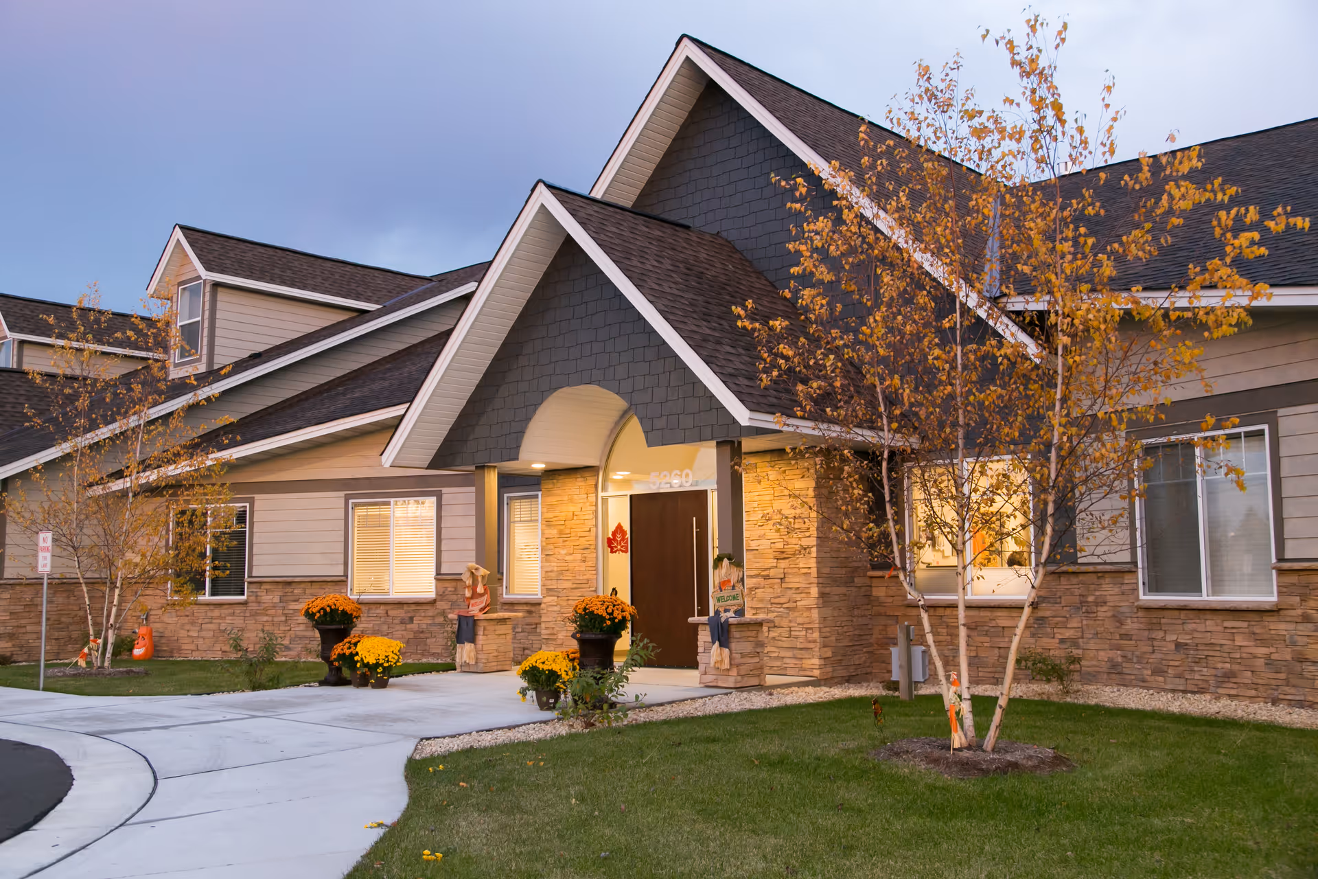 Exterior front entrance of a single-story residential building with a covered porch, stone accents, potted chrysanthemums, and autumn trees.