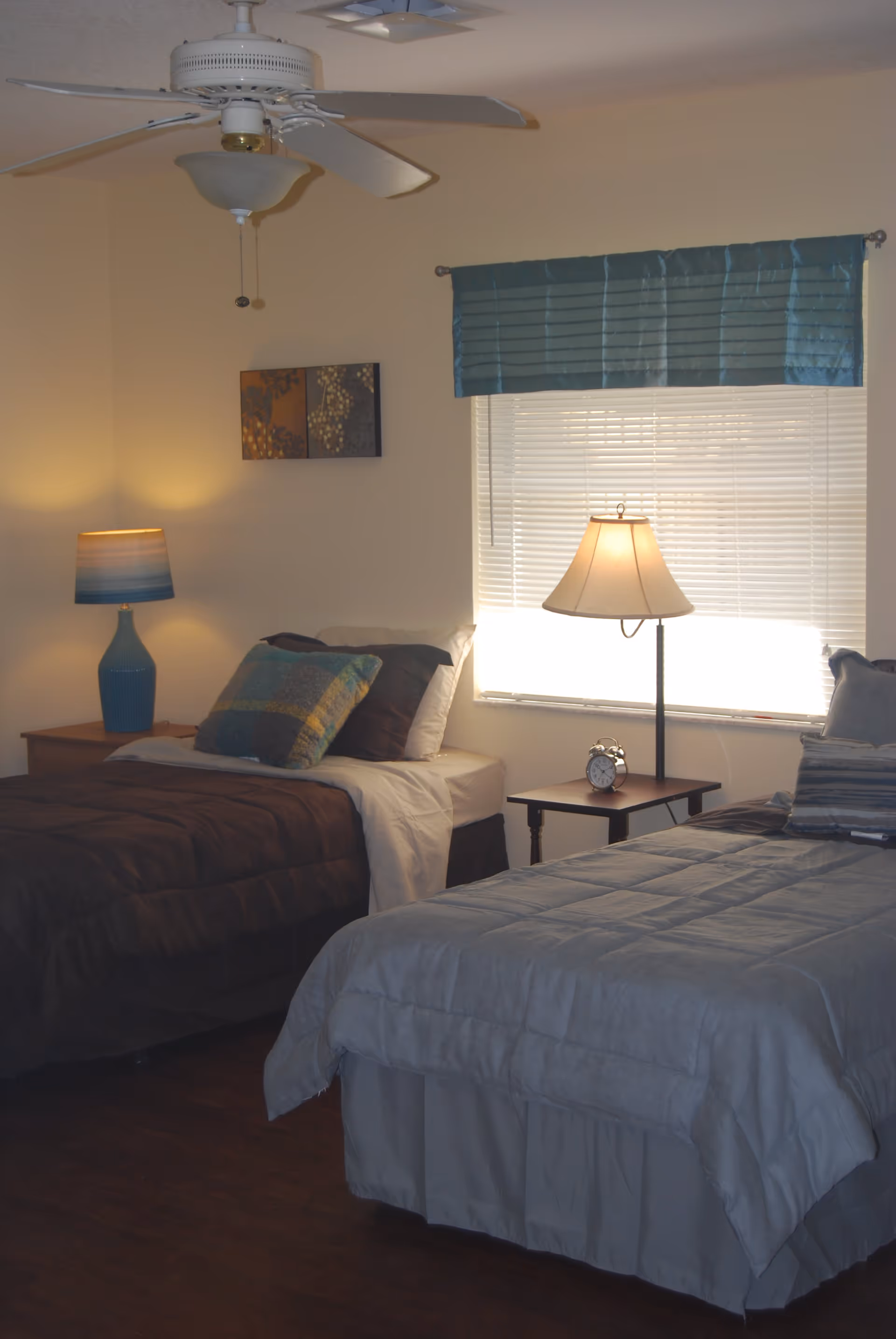Two twin beds in a softly lit bedroom with bedside tables, lamps, a window with blinds, and a ceiling fan.