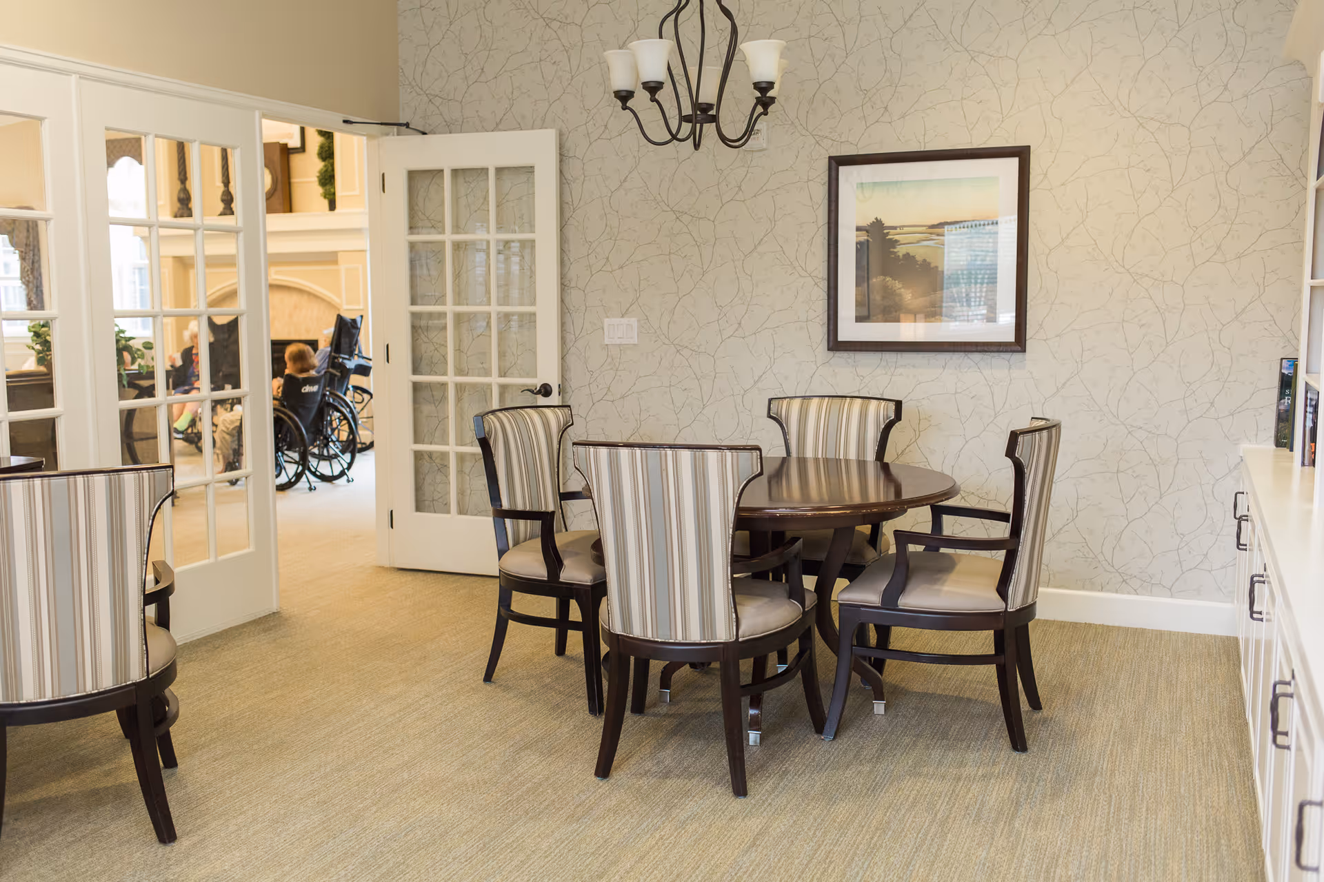 A dining area with a round wooden table surrounded by five striped upholstered chairs. The room has light-colored wallpaper with a subtle pattern, a chandelier hanging from the ceiling, and a framed picture on the wall. Through glass-paneled double doors, a living room area is visible with people, including one person in a wheelchair.