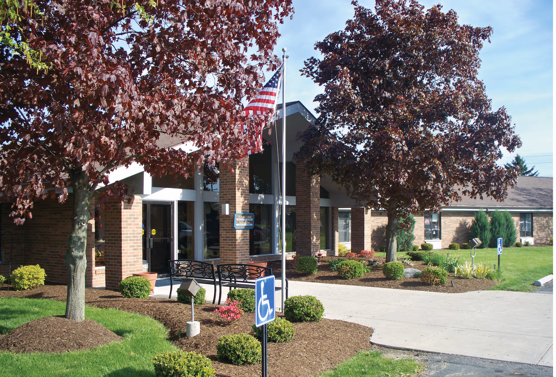 Entrance of Delaware Court Healthcare Center showing a brick building with large windows, two trees with red leaves, a flagpole with an American flag, a bench, and a sidewalk with handicap parking signs.