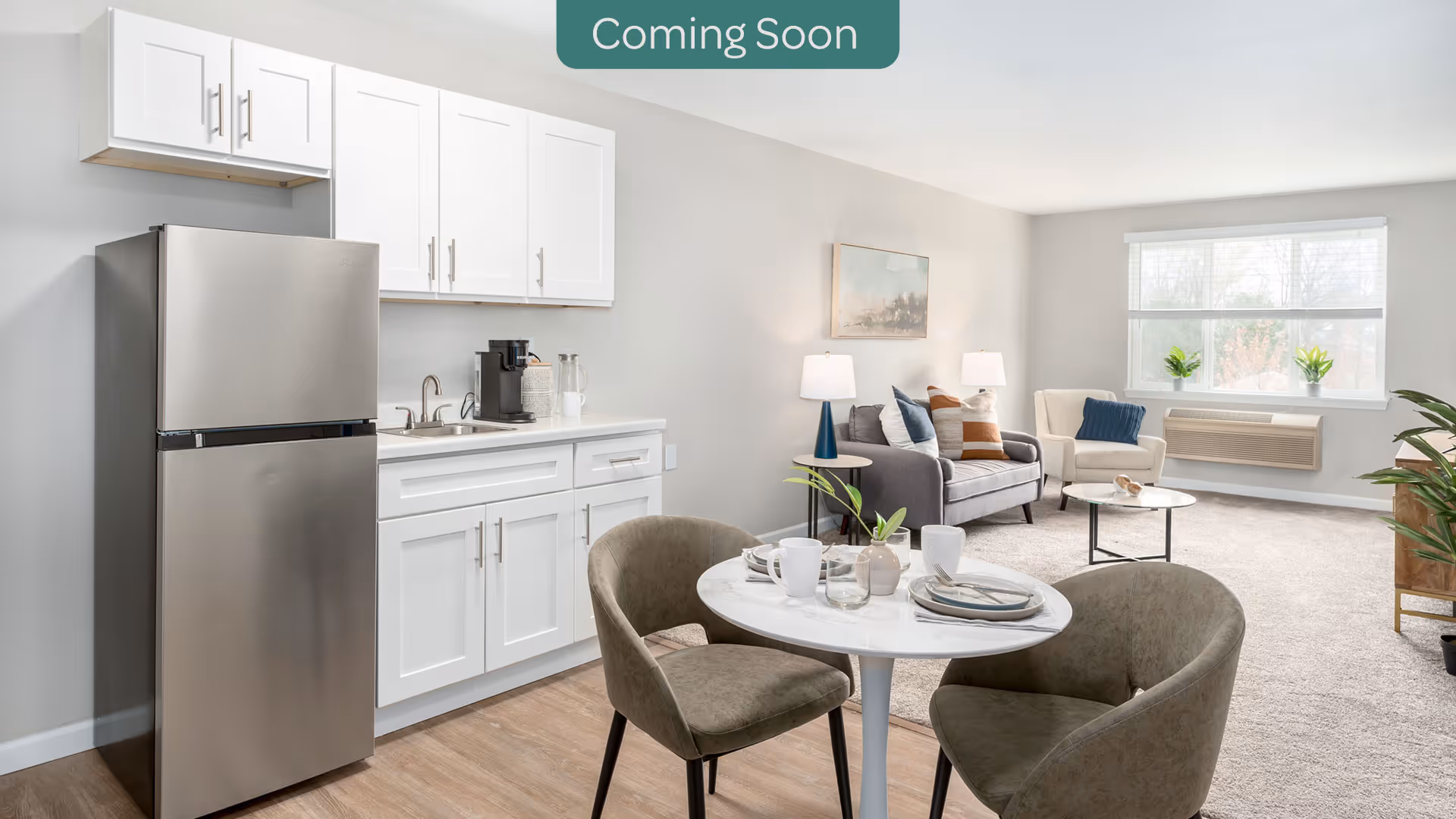 A bright and modern open-concept living space featuring a small kitchen area with white cabinets, a stainless steel refrigerator, and a coffee maker on the counter. In the foreground, there is a round dining table set with plates, cups, and a small plant, surrounded by two upholstered chairs. The living area in the background has a gray sofa with colorful pillows, a beige armchair, a round coffee table, two table lamps, and a large window letting in natural light.