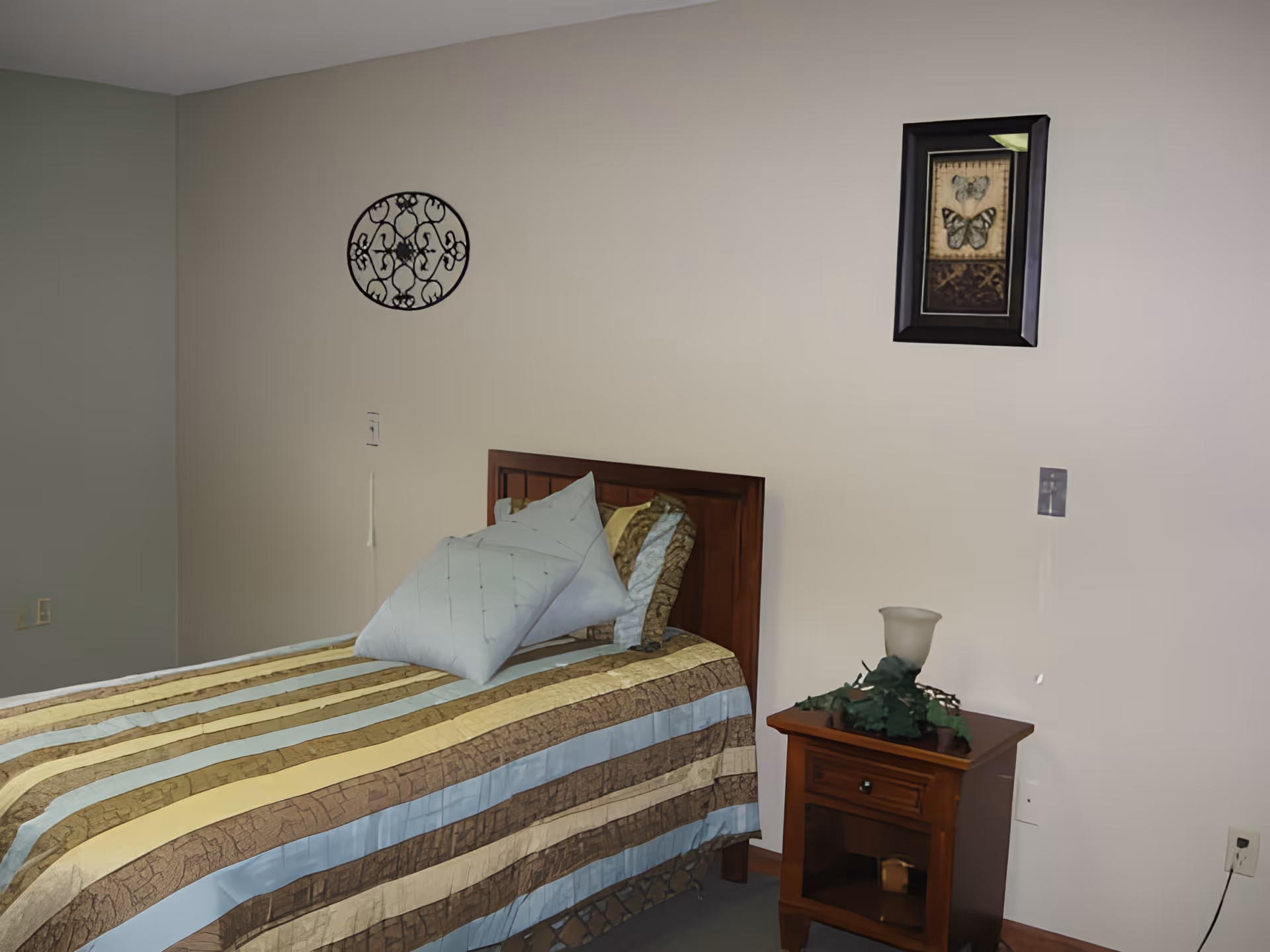 A simple bedroom with a single bed covered in a striped bedspread in shades of brown, yellow, and blue. Two pillows are placed on the bed. Next to the bed is a wooden nightstand with a decorative plant and a frosted glass lamp. The walls are plain with a round metal wall decoration and a framed butterfly picture hanging above the bed.