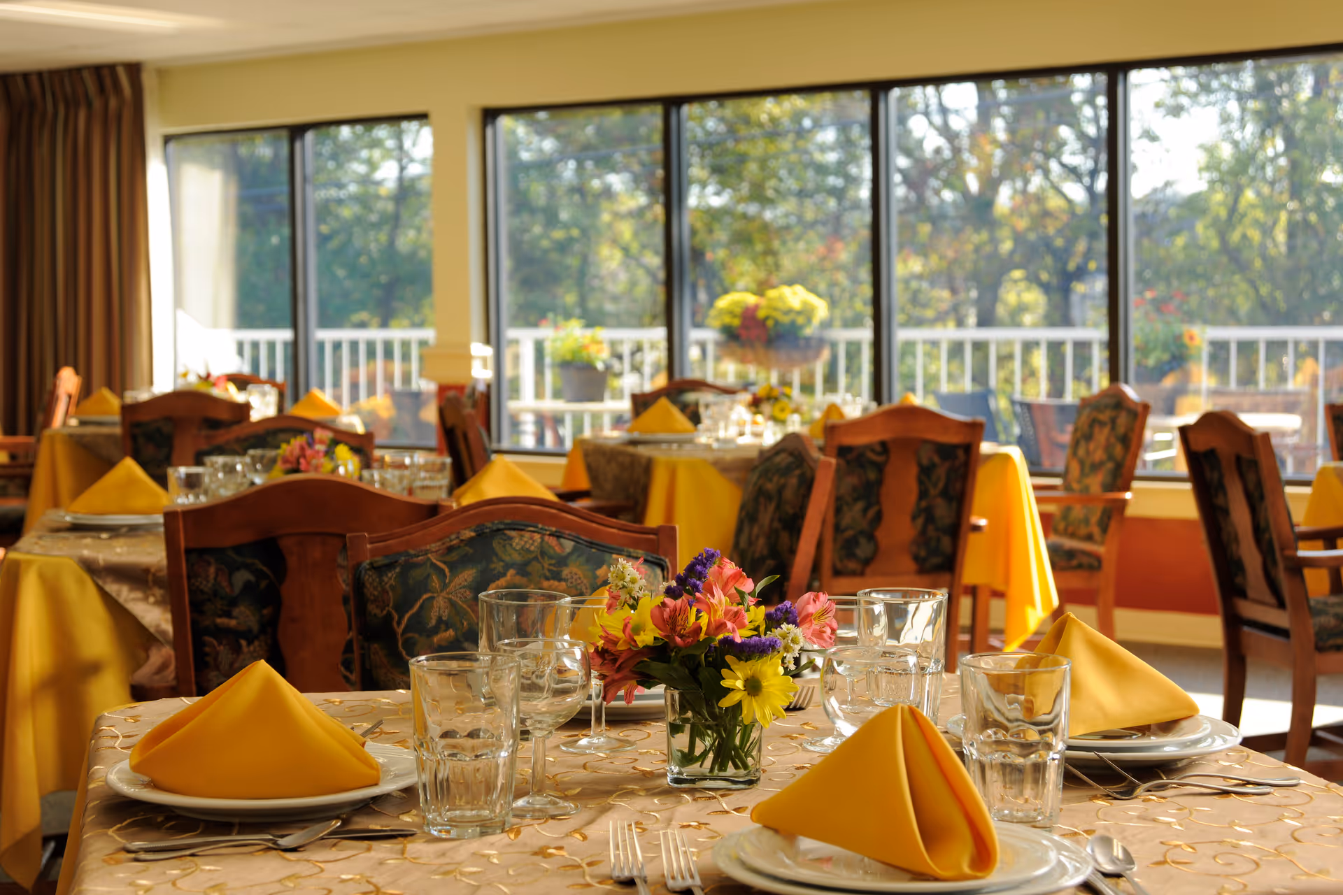 Dining room with tables set for a meal, featuring yellow folded napkins, glassware, plates, silverware, and floral centerpieces. Large windows in the background show an outdoor view with greenery and flowers.