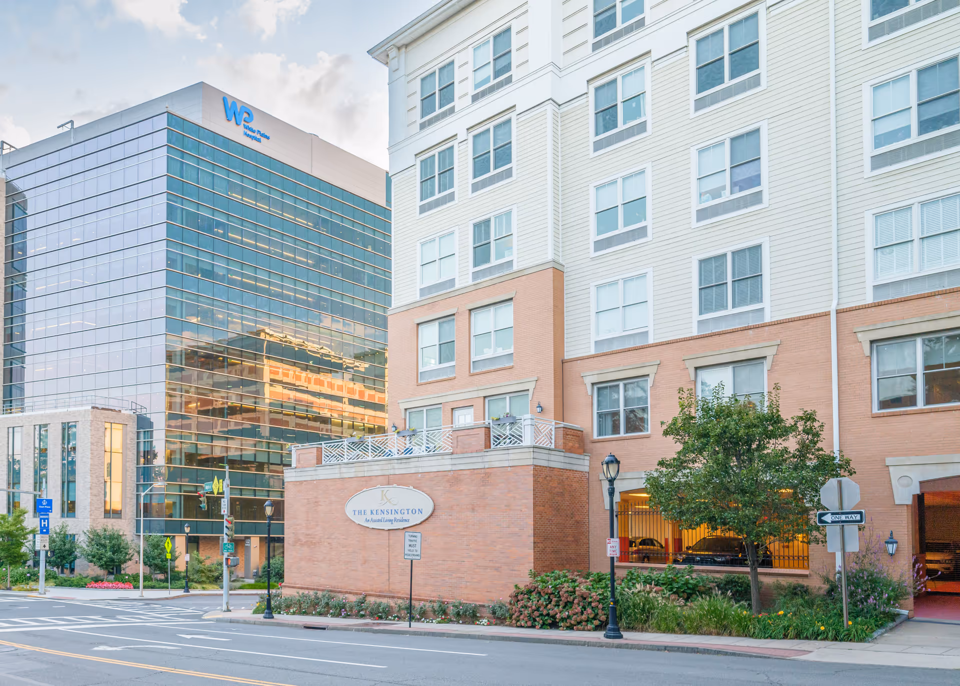 Exterior view of The Kensington White Plains building, an assisted living residence, showing a multi-story structure with brick and light-colored siding. The building is situated on a street corner with street signs, a tree, and some landscaping. In the background, there is a modern glass building with the sign 'White Plains Hospital'.
