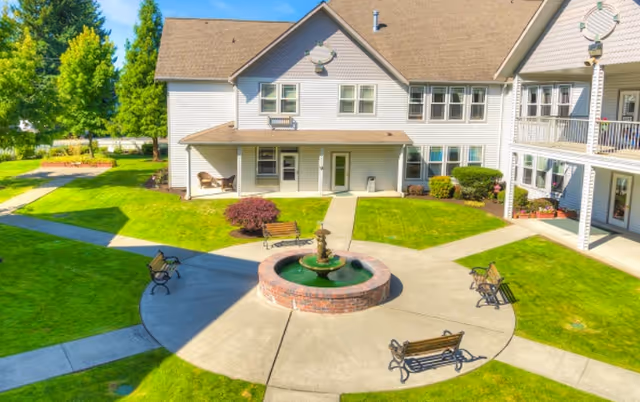 Outdoor courtyard area of a senior living facility with a circular brick fountain in the center, surrounded by four benches on a concrete pathway. The courtyard is bordered by green grass and pathways leading to a two-story building with white siding and multiple windows. Trees and shrubs are visible in the background under a clear blue sky.