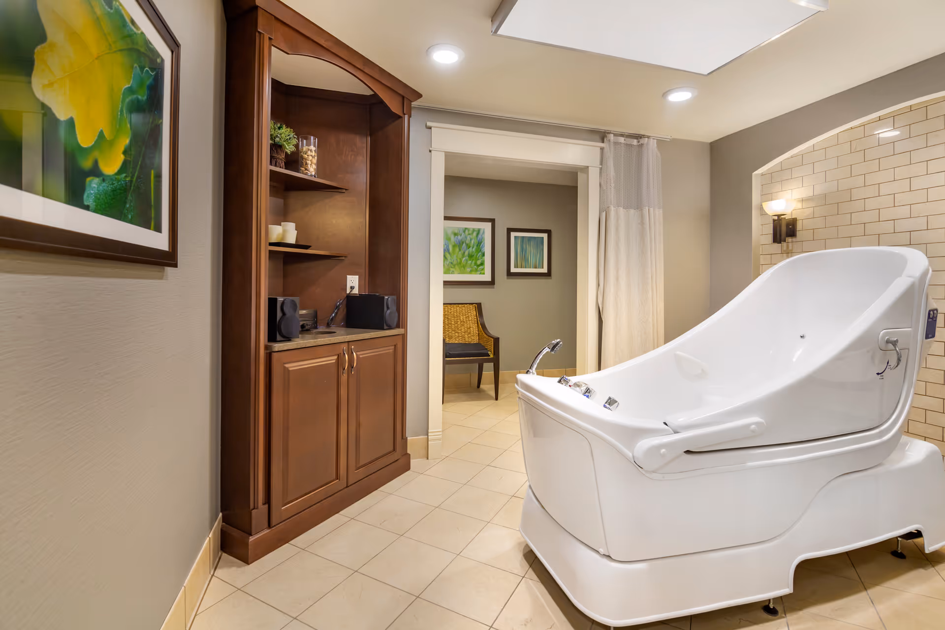 A modern bathing room featuring a large white therapeutic bath tub with controls on the side, beige tiled floor and walls, a wooden cabinet with shelves holding decorative items, and framed artwork on the walls. There is also a chair visible through an open doorway in an adjacent room.