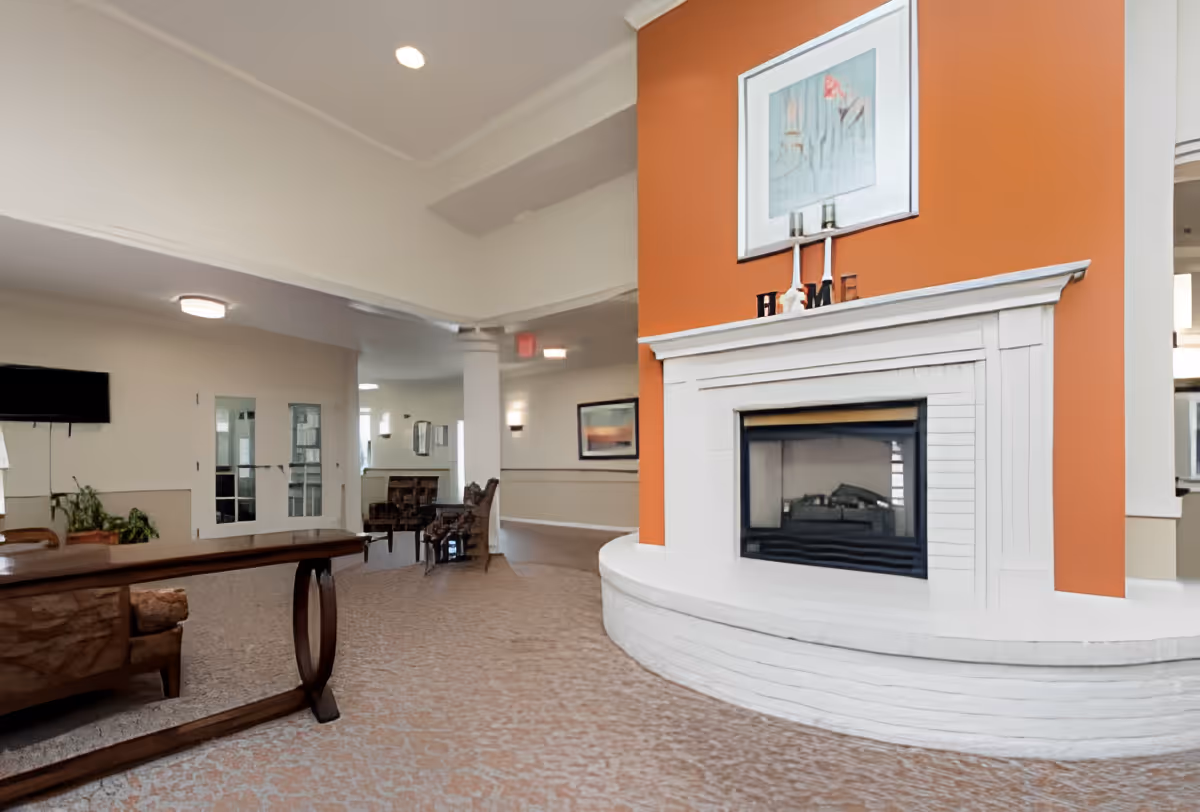 Spacious senior living common area with a large white fireplace against an orange accent wall, seating, and a TV.