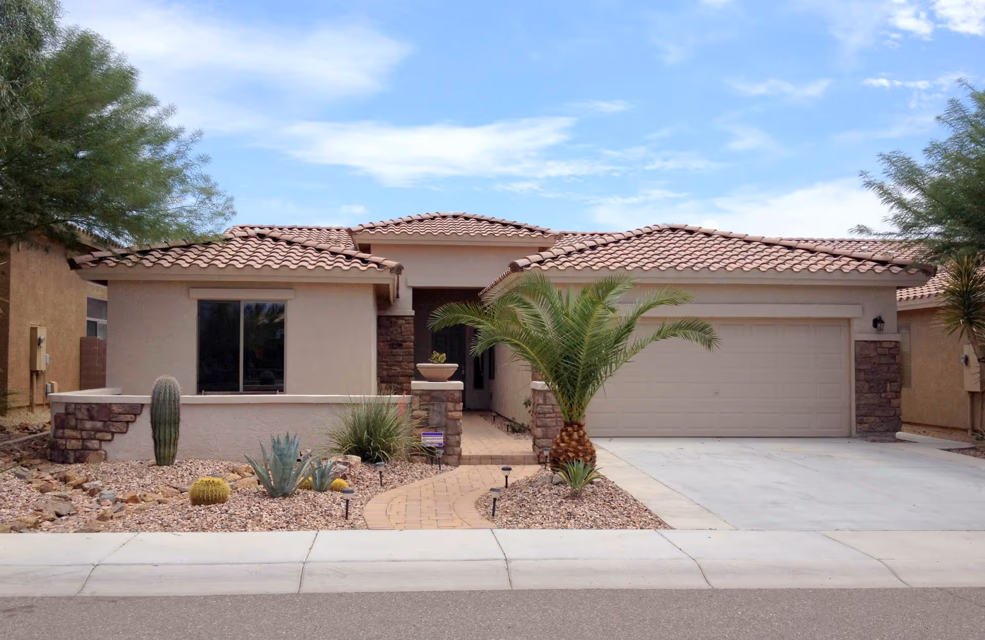 Single-story stucco house with a tiled roof, attached two-car garage, and desert landscaping with a palm and cacti.