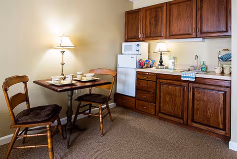 Small kitchen area with wooden cabinets, a white mini refrigerator, microwave, and countertop with a lamp, dish soap, and a towel. Next to the kitchen is a small dining table set for two with wooden chairs and a table lamp on the floor.