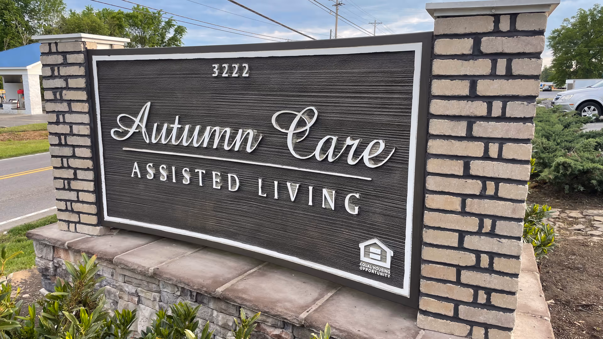 A large outdoor sign for Autumn Care Assisted Living with the address number 3222 displayed above the name. The sign is mounted on a stone and brick base with some greenery around it. A road and some parked cars are visible in the background.