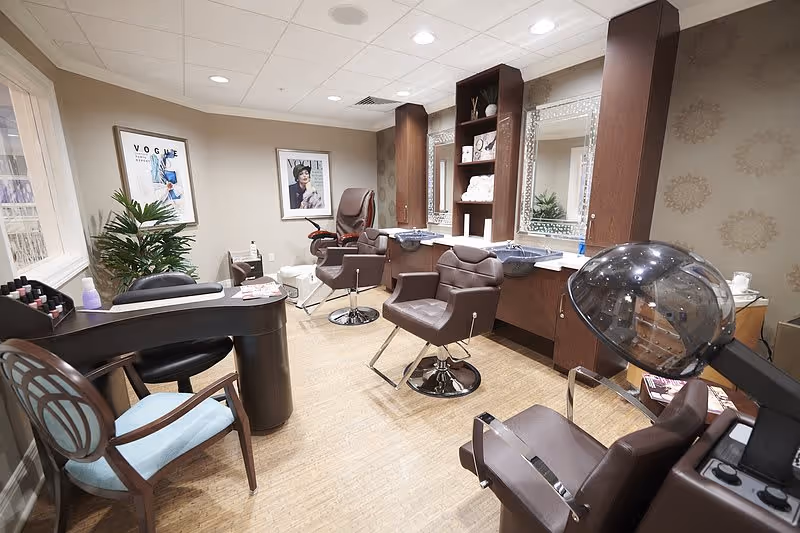 Interior of a senior living facility salon with multiple brown salon chairs, a hair dryer, two large mirrors, shelves with towels and hair products, a manicure table with chairs, and framed Vogue magazine covers on the wall.