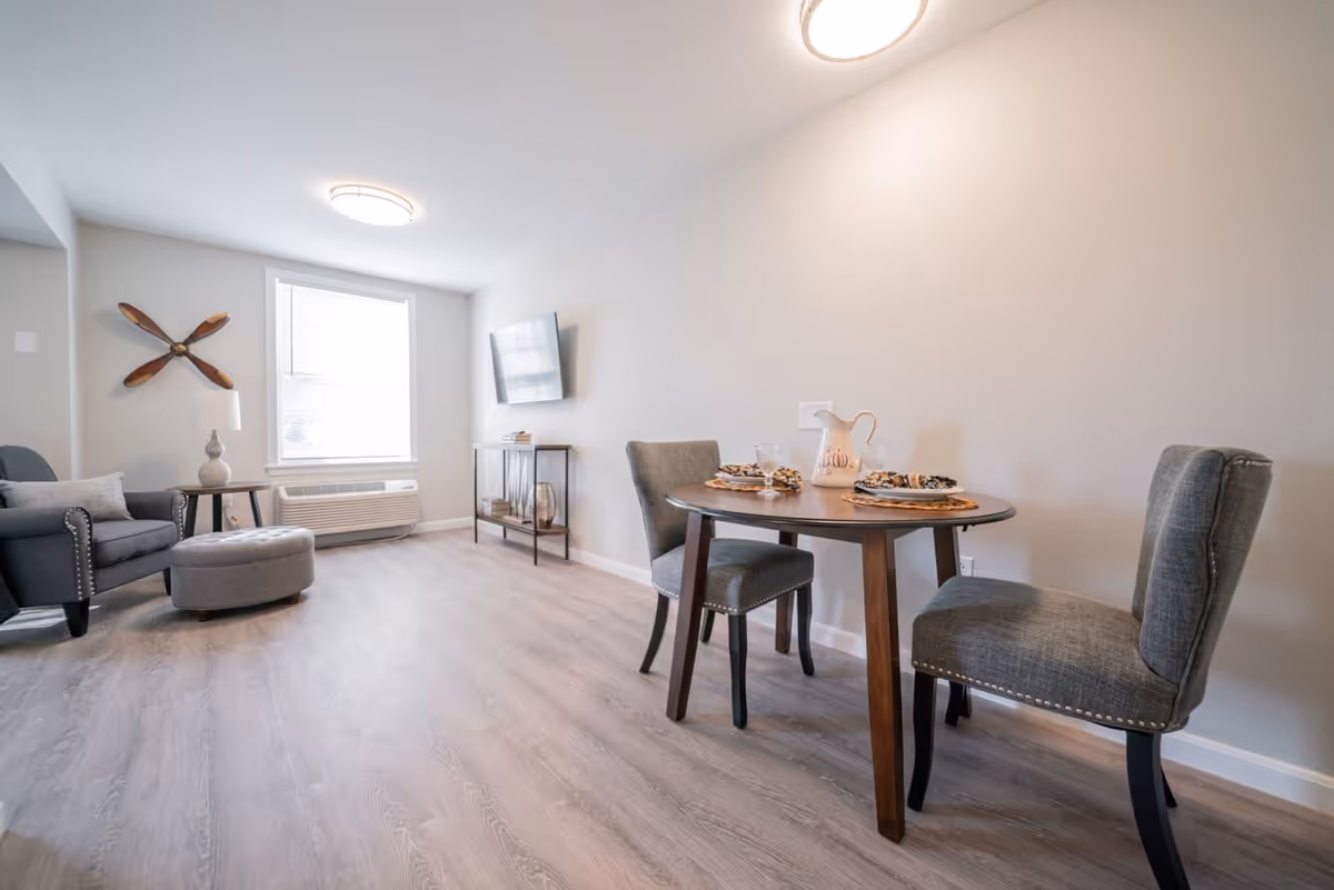 A bright and modern living area with light wood flooring, a small round dining table set for two with gray upholstered chairs, a gray armchair with a matching ottoman, a side table with a lamp, a wall-mounted flat-screen TV, and a decorative wooden propeller on the wall near a large window.