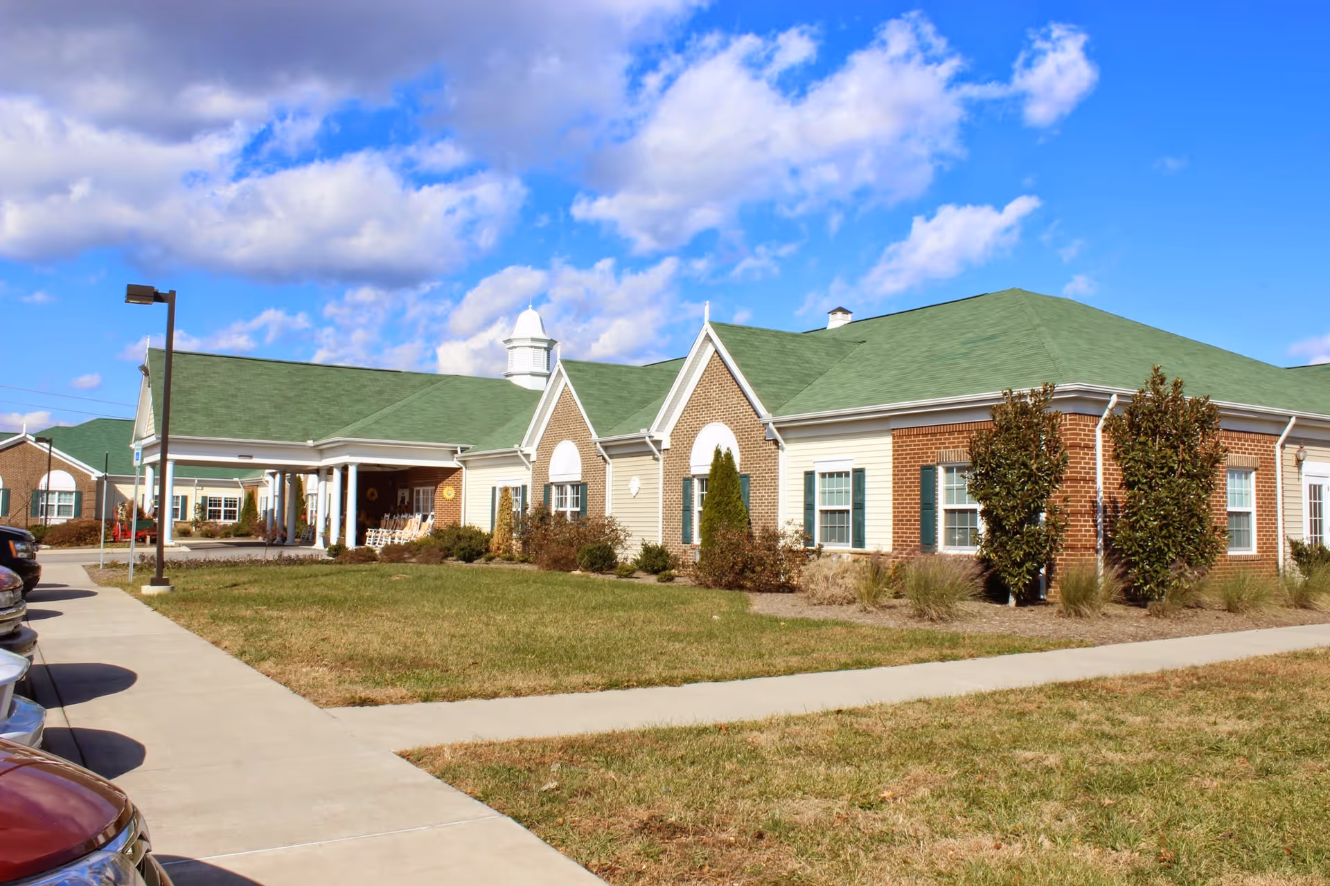 Exterior view of Meadow View Senior Living facility showing a single-story building with green roofs, brick and beige siding, surrounded by a lawn and a sidewalk. Several cars are parked along the driveway on the left side under a partly cloudy blue sky.