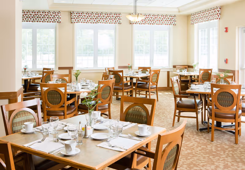 Bright dining room with multiple wooden tables and chairs set with white plates, cups, glasses, and silverware. The room has large windows with patterned valances allowing natural light to fill the space. Small flower arrangements are placed on each table.
